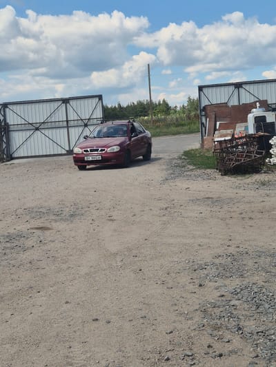 A burgundy Daewoo Lanos sedan with license plate BA 1884 AK is parked on a gravel lot. The car is facing right and slightly angled towards the viewer. Behind the car, a large white corrugated metal gate is visible on the left side of the image. To the right of the car, there is another building or structure with corrugated metal siding, and a pile of rusty metal farm equipment is stacked in front of it. The environment appears to be an outdoor industrial or agricultural area, possibly a junkyard or storage facility, under a partly cloudy sky. The location is identified as Pidhaitsi, Ukraine.
