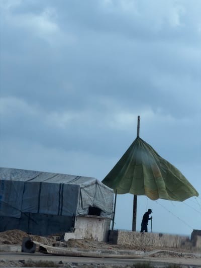 In the city of Nuseirat, Palestinian Territories, under a cloudy sky, a man stands silhouetted under a large, olive-green tarp, held aloft by a central pole. The tarp is stretched out like an umbrella, casting a shadow over the man. He is wearing a dark hat and holding a long stick. To the left of the man and the tarp is a large, temporary structure, possibly a tent, covered in a grey tarp with vertical black stripes. This structure appears to be on a raised concrete platform. The foreground shows a debris-strewn area with piles of sand and what looks like discarded construction materials. The overall scene suggests a temporary settlement or construction site.