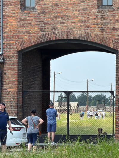 The image depicts a group of people near a chain-link fence in Brzezinka, Poland. The scene appears to be a historical site, possibly a former camp, indicated by the presence of old brick buildings, a chain-link fence, and what seem to be remnants of structures in the background. The sky is clear and bright, suggesting it is daytime, likely on a sunny day.
Several individuals are visible in the foreground and mid-ground. One person, wearing a blue patterned shirt and blue shorts, stands near the fence. Further back, two people are seen, one in a gray t-shirt and blue leggings, and another in a blue t-shirt and shorts, looking towards the right of the frame. In the background, through the fence, a group of people in white shirts are gathered, possibly on a guided tour. The overall mood seems somber, consistent with a memorial site.
Notable details include the texture of the old brickwork, the open sky, and the presence of lush green grass and foliage to the right of the frame. A white car is parked on the right side of the image. There is a license plate on the car with visible text, but it is illegible due to its size and resolution. The scene is set outdoors, with the bright sky and natural vegetation indicating it is likely spring or summer.