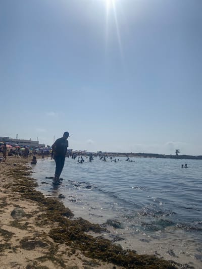 The image depicts a bustling beach scene in Harhoura, Morocco, on a sunny day. The foreground features a sandy beach strewn with green seaweed, transitioning into clear, shallow water. Several people are visible wading and swimming in the sea. In the mid-ground, a man wearing a dark t-shirt with a "NIKE" logo and a baseball cap stands at the water's edge, facing away from the camera. The background reveals a crowded beach lined with colorful umbrellas and people relaxing on the sand. Further in the distance, buildings and a lifeguard tower are visible. The sky is bright blue with a strong sun flare in the upper left corner.