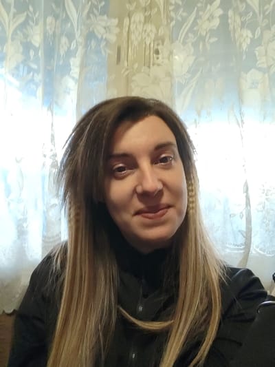 The image displays an adult female captured in a head-and-shoulders view, positioned indoors within the city of Opaka, Bulgaria. The subject is centered, facing the camera directly with a subtle, neutral smile. She possesses long, multi-toned hair, appearing light brown to blonde with darker sections near the scalp, and a distinct crimped texture is visible on the hair strand descending along the viewer's left side of her face. She is dressed in a dark-colored, long-sleeved garment, likely a collared shirt or blouse, with several buttons visible on the front placket. The background consists of a window covered by a light-toned, translucent lace curtain featuring a repeating floral pattern, through which bright ambient light is perceived.