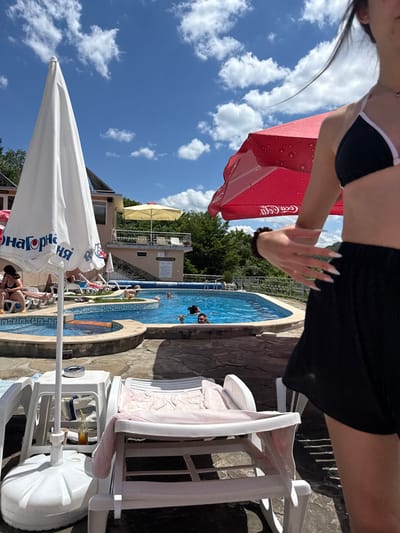 This is an outdoor, eye-level, medium shot photograph taken at the Shipkovo mineral pools in Bulgaria. The scene captures a sunny day with a bright blue sky filled with white, fluffy clouds. In the foreground, a person's torso and legs, wearing a black bikini top and black shorts, are partially visible on the right side of the frame. Their hand, with long, pointed fingernails, is extended outwards, partially obscuring a red umbrella.
Behind the person, a swimming pool complex is visible. A large white umbrella stands prominently in the left foreground, displaying Cyrillic text that appears to say "Здраве От Народна" (Health from the People). Adjacent to the white umbrella, a white plastic lounge chair holds a pink towel and a partially visible pink blanket. Another white lounge chair is behind it.
The swimming pool itself is oval-shaped with blue water and a mosaic tile border. Several people are in the pool, including two men and a woman, one of whom is looking towards the camera. Another person is visible in the background sitting on a lounge chair. The pool area is surrounded by stone paving.
In the background, a light-colored building with a balcony is visible, and behind that, dense green trees line a hillside. A smaller yellow umbrella is seen on the balcony of the building. The overall atmosphere suggests a relaxed, summery day at a recreational facility.