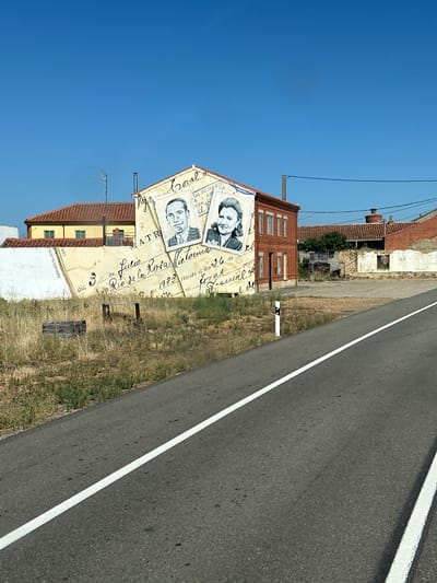 In Toral de Fondo, Spain, a mural is painted on the side of a building. The mural depicts a large, yellowed envelope with cursive writing and two portraits of individuals. The building itself is red brick with white-trimmed windows and a sloping tiled roof. The mural covers the side of the building, extending upwards to the roofline. Adjacent to this building is another structure with a red tiled roof, partially obscured. The foreground features a paved road with a white line marking its center, bordered by dry, grassy vegetation. The sky above is a clear, vibrant blue.