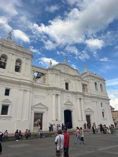This image shows the exterior of the León Cathedral in León, Nicaragua. The cathedral is a large, white neoclassical building with a prominent facade featuring several arches, columns, and decorative elements. Two bell towers are visible, one on the left with three bells and another on the right with arched openings. Statues are present on the facade, including a figure with a halo at the top center and two statues supporting a bell in an upper archway.

In front of the cathedral, a large stone lion statue stands to the left of the central entrance. People are gathered in the plaza in front of the building. Some are sitting on a low wall to the left, while others are standing and walking around. A group of people, some in formal attire such as dresses and suits, are gathered near the entrances, suggesting an event or ceremony is taking place or has recently concluded. The sky is blue with scattered white clouds.