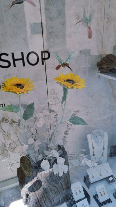 The image shows a shop window display in Rupit, Spain. The window features several decorative items, including artificial sunflowers, decorative hummingbirds made of what appears to be glass or plastic, and various jewelry pieces displayed on white stands. There is also a rustic wooden planter holding dried plants and a piece of driftwood on a shelf. The background appears to be a textured wall with the word "SHOP" visible, indicating it is a retail establishment. Sunlight is casting shadows on the scene, suggesting it is daytime. No people are visible in the image.