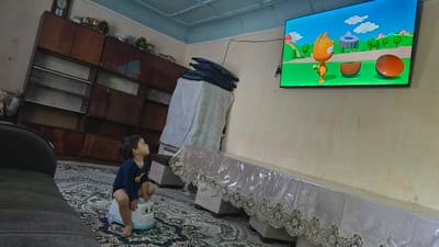 In a room with a patterned carpet, a young child sits on a toy car, facing a television screen. The child, wearing a dark blue long-sleeved shirt and no pants, looks intently at the animated characters on the screen. The television displays a cartoon with a bright green landscape, featuring a small red barn, trees, and whimsical structures. In the foreground, the child is partially obscured by a dark grey sofa. Behind the child, a large wooden cabinet with glass doors and open shelves stands against the wall. Decorative items, including vases and greenery, are visible on top of the cabinet. To the right of the child, a long, low surface is covered with a decorative cloth. The room appears to be illuminated by indoor lighting. The overall scene suggests a child engaged in watching cartoons in a home environment.