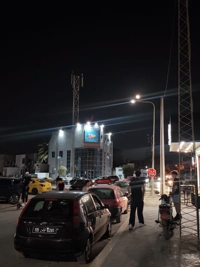 The image depicts a street scene at night in Ariana, Tunisia. A modern, multi-story building with a prominent illuminated sign featuring a logo with the letters "TT" is visible in the background. The building's facade is illuminated by bright lights. Several cars are parked and driving on the street, including a black Ford Fiesta with a Tunisian license plate that reads "6281 تونس 115". A yellow taxi is also present.
Several individuals are present. One person is standing on a kick scooter, dressed in dark clothing and a white cap. Another person, also in dark clothing, is walking away from the camera. Two other individuals are standing near a motorcycle on the sidewalk. A red stop sign is mounted on a pole. Streetlights illuminate the scene, casting long shadows on the pavement. A cell tower is visible in the upper portion of the image. The environment appears to be urban, with sidewalks, streetlights, and buildings.