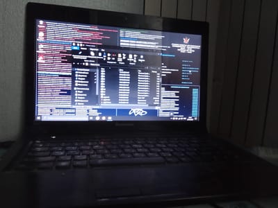 This is a close-up, dimly lit shot of a Lenovo laptop placed on a desk. The laptop screen is on, displaying a complex interface with multiple windows and a file explorer. The screen shows a mix of Russian text and what appears to be code or system information.
The primary subject is the laptop itself, along with its illuminated screen. The screen displays what looks like a work-related application or system status, with various panes and lists of data. One prominent window is a file explorer, showing file names, dates, and sizes. There's also a graphic resembling a logo on the screen, possibly for an organization.
The scene is dark, suggesting it's either nighttime or the room is otherwise dimly lit. The background consists of vertical blinds or shutters, indicating an indoor setting. There are no people visible in the frame.
Visible text on the screen includes: "ГЛАВНЫЕ РАБОТЫ ОРГАНИЗАЦИИ", "ПЕРЕРЫВ НА ОБЕД", "13:00 - 14:00", "ОСНОВНАЯ ОБЛАСТЬ", and numerous other lines of text in Russian, some of which appear to be file paths or data entries. The file explorer window has column headers like "Имя", "Дата изменения", and "Тип". The brand name "Lenovo" is visible on the laptop's bezel below the screen.
The overall impression is one of focused work or activity on the computer in a private, perhaps office or home, environment. The low light and the content on the screen hint at a serious or professional task being undertaken. The location context of Kalachinsk, Russia, suggests the text on the screen is likely in Russian.