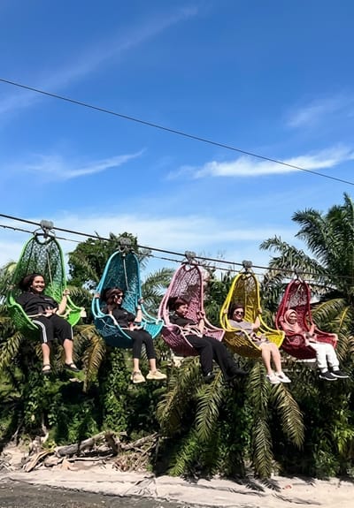 Five individuals are seated in colorful, hanging basket-style swings suspended from a wire, likely a zipline attraction, against a backdrop of lush green tropical foliage and a bright blue sky with scattered white clouds. The swings are arranged in a row and are colored green, blue, pink, yellow, and red from left to right. Each person is wearing casual clothing and appears to be enjoying the ride, with some smiling and looking at the camera. The scene suggests an outdoor recreational activity in a natural setting. The location is identified as Bintang Meriah, Indonesia.