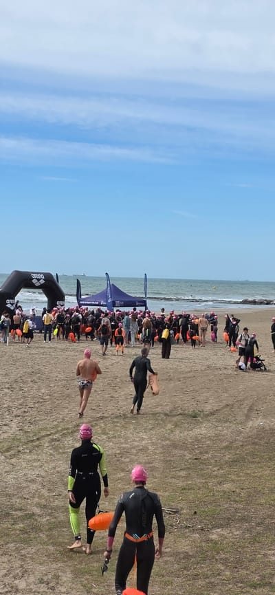 The image depicts a triathlon event on the beach in Benicàssim, Spain. Swimmers are exiting the water and running onto the sand, some wearing wetsuits, pink swimming caps, and carrying orange buoys. An "Arena" branded inflatable archway marks the transition area. Spectators and event staff are visible along the beach. The sky is blue with scattered clouds, and the sea is calm.