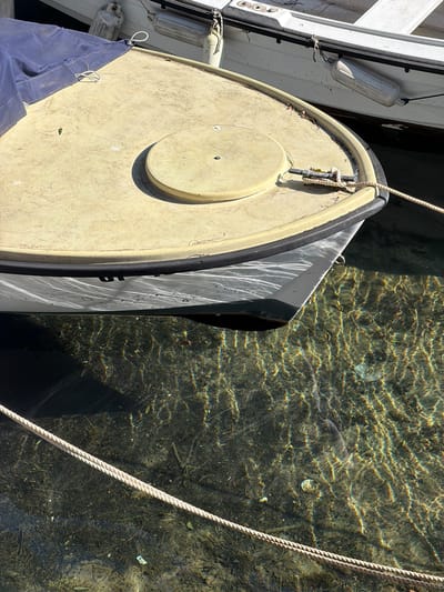 The image captures a close-up of the bow of a small boat, likely a dinghy, moored in the clear waters of Grad Opatija, Croatia. The boat features a rounded bow with a light-colored deck and a darker hull. Ropes secure the boat. The water reveals a sandy or pebbly bottom with visible details. Light refracts on the surface, creating shimmering patterns. Another boat is visible to the left, alongside the dinghy.
