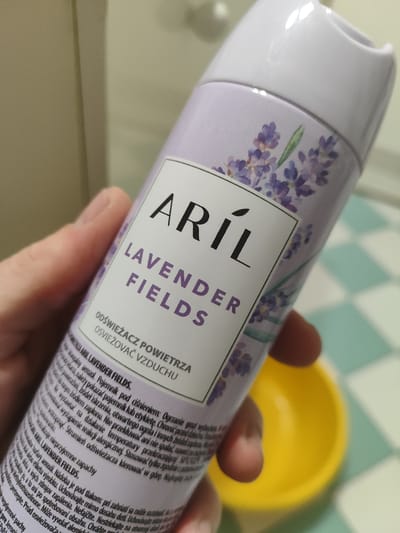 A person's hand is holding a can of "ARIL LAVENDER FIELDS" air freshener. The can is predominantly lavender in color with a white label featuring purple lettering and a floral design. The label contains text in both Polish and Czech, describing the product as an air freshener. The background shows a muted, light-colored wall or surface, with a portion of a tiled floor visible in the lower right corner. A bright yellow bowl is partially visible in the foreground, to the right of the can. The lighting is soft, suggesting an indoor environment. There is no discernible information in the image that would suggest the location is specifically in the city of Łomianki, Poland.