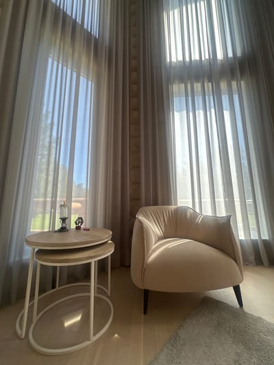 A partially visible armchair sits adjacent to a set of stacking tables in front of a window covered by sheer curtains. The scene is indoors. A small glass lamp and a few small objects are sitting on top of the upper table. The scene is likely set within a home. No people are visible. The location in Areyskoye, Russia, is not verifiable.