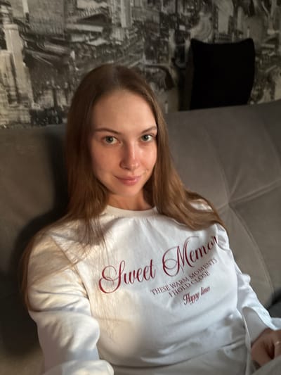 A young woman with long, auburn hair is seated on a grey sofa. She is looking towards the camera, with a slight smile. She is wearing a white long-sleeved shirt with red text that reads "Sweet Memories". The background appears to be a wall with a black and white city-themed wallpaper. The overall scene suggests an indoor setting, likely a home, and based on the provided metadata, the location is Novaya Ivanovka, Russia.