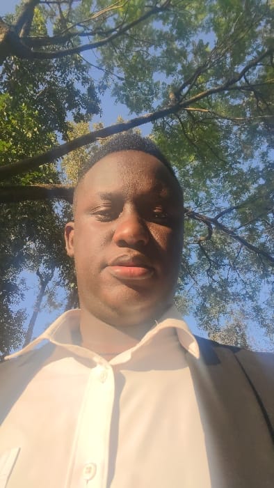 A person poses in front of tree branches and blue sky. The person wears a light-colored collared shirt. The face is angled towards the camera, with closed eyes and sunlit skin. The background features lush green leaves and branches silhouetted against the sky. The camera angle is low, looking up at the person and the canopy above.