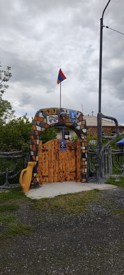 A wooden gate with a brick archway is situated in Dashtadem, Armenia. The archway is decorated with depictions of animals and portraits of a man. The Armenian flag is mounted on top of the archway. The gate is flanked by a yellow jar and a tree-like structure. The ground is covered with gravel and grass. A streetlight is visible in the background. A house can be seen through the gate. The sky is overcast.