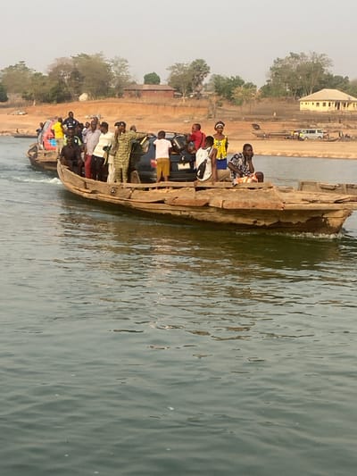 A wooden boat crowded with people, including one in military uniform, and a dark car is crossing a body of water. The aged boat is made of rough wood planks. Reflections ripple on the water's surface. In the background, a tan landscape features a building and trees under a hazy sky. The scene appears to be a local ferry transporting people and vehicles.