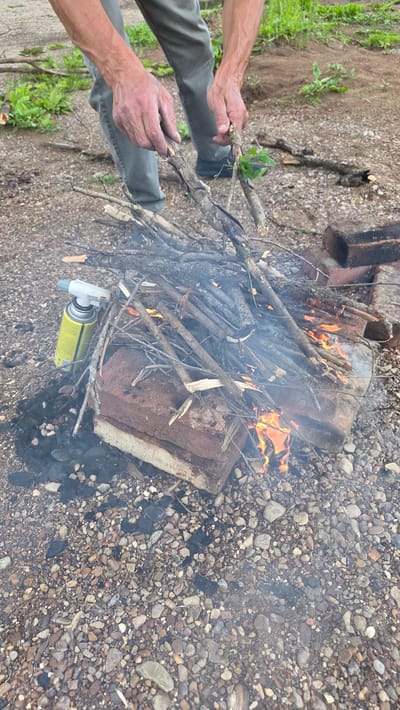 In Дарьино, Russia, a person's hands are adding dry twigs to a small bonfire. The fire is situated on a patch of gravel, with a few pieces of brick serving as a makeshift fire pit. A yellow butane torch is placed beside the fire pit. The background shows dry soil and sparse green vegetation. The overall scene depicts the process of starting an outdoor fire.