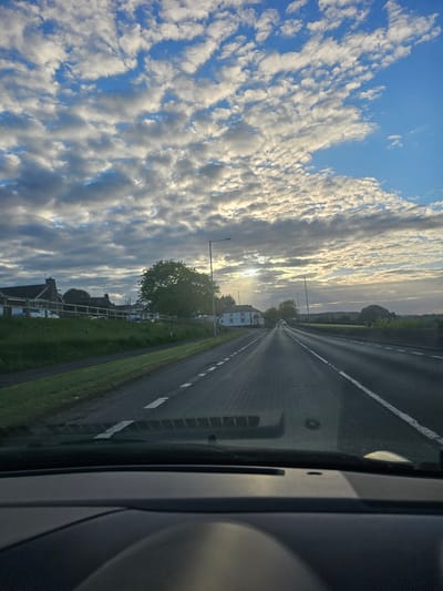 The image is taken from inside a vehicle, possibly a car, looking out onto a road in what appears to be a suburban or rural area. The road is a straight, two-lane road with white lane markings and a green grassy shoulder on the left side. On the left side of the road, there are houses and streetlights. In the distance, the road appears to lead towards the sun, which is partially obscured by clouds. The sky is filled with a mix of blue and clouds. The interior of the vehicle is visible on the right side of the image. The city is not identifiable.
