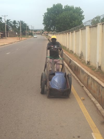 The image shows a man pushing a handcart along a paved road in Kanshio, Nigeria, located at latitude 7.683 and longitude 8.537. The man is wearing a yellow hat and a dark shirt. The handcart is rusty and has two wheels, one of which is larger than the other. A blue, perforated, round object is in the cart. The man is facing the camera, walking toward the left. There are a few palm trees and buildings along the street and a long beige wall lined with barbed wire along the right side of the road. The road is mostly empty except for a small white car in the distance. There is a yellow line separating the right lane of the road from the sidewalk. A small, light-colored rock lies in the right lane. The sky is overcast and gray. The man is walking toward the left. The image was likely taken by a person standing on the sidewalk to the right.