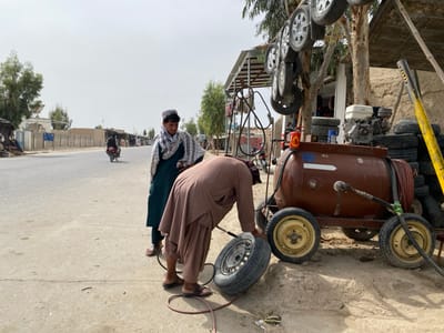 The image shows a man in a brown robe bending over to work on a tire next to a brown air compressor. The air compressor is attached to a cart and has a red tank, a gray handle, and a black hose leading from it.  Behind him, a man wearing a green robe and a patterned scarf is standing on a street.  There is a motorbike on the street to the left of the two men and a stack of tires behind them, leaning against a tree.  The scene is located in Panjwai, Afghanistan, at latitude 31.542 and longitude 65.463.  There is a building in the background, and the ground is made of packed dirt.