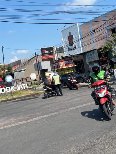 The image depicts a street scene in Entalsewu, Indonesia. A red scooter with a rider wearing a green jacket is in the foreground, moving from left to right. The scooter's license plate reads "W 4307 NBW". In the background, there are several buildings, including one with a sign that says "haus!". A large white sign with the text "DOK JATI" is visible on the left side of the image. There are also other people on motorbikes and walking on the street. The sky is blue with some clouds, and there are power lines overhead. The street appears to be paved, and there is some vegetation on the sides. The overall impression is a bustling street in a tropical or subtropical urban environment.