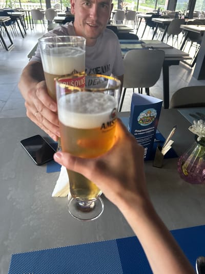 Two people are toasting with glasses of beer at a table in a restaurant. One person, a male with short hair and a light-colored t-shirt, is holding a glass in the foreground, while another person's hands are visible holding a second glass in front of him. The table is gray and set with blue placemats. A black smartphone lies on the table. In the background, there are other tables and chairs, suggesting a dining area. A blue triangular brochure with text in Russian and an illustration of mountains and a sun is visible on the table, along with a pen holder with pens and a small vase with a white flower. The scene appears to be indoors, with natural light coming from large windows, and an outdoor view of greenery visible beyond them. The location is identified as a restaurant in Danilovka (Altai Krai), Russia.