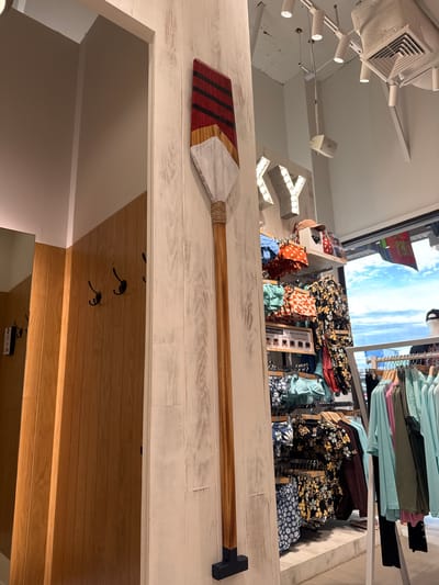 The image appears to be taken inside a retail store, possibly a surf or beachwear shop, located in Krabi, Thailand. A decorative wooden paddle with red, white, and natural wood sections is mounted vertically on a white-washed wooden wall in the foreground. To the left, there's a glimpse of a changing room with wooden paneling and black coat hooks. In the background, clothing is displayed on racks, including swimwear with floral and patterned prints, and various t-shirts and tops in pastel colors. The store features bright overhead lighting and a large window in the back showing a cloudy sky and some tropical vegetation. The overall ambiance suggests a relaxed, beach-themed retail environment. There are no people visible in the image.