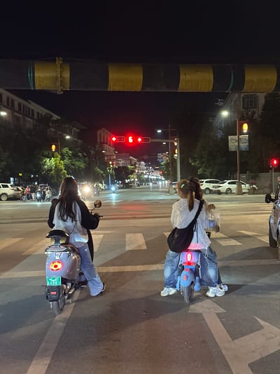 At night, on a street in Eshan Yi Autonomous County, China, two individuals, likely women, are riding on electric scooters at a crosswalk. One scooter, colored gray, has a license plate with the characters "A4881" and is occupied by a person wearing blue jeans and a black and white jacket. The other scooter, colored blue, is ridden by a person in blue jeans and a white top, with another person behind them. Both scooters have their taillights illuminated. In the background, traffic lights show red, indicating a stop. Several vehicles, including cars and motorcycles, are present on the street. Buildings with illuminated windows line the sides of the road, and trees are visible. A yellow and black striped structure, possibly part of a traffic signal or overpass, is suspended above the street.