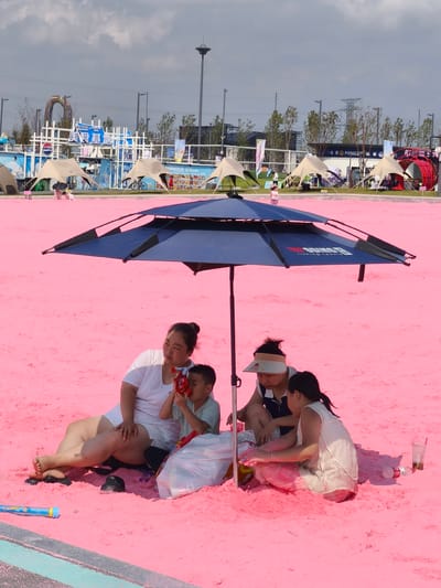A family of four is relaxing on a pink sand beach under a large blue umbrella. The family consists of a woman sitting on the left, a young boy sitting in front of her, and a woman and a girl sitting on the right. The woman on the left is wearing a white t-shirt and white shorts. The boy is wearing a light blue t-shirt and shorts. The woman on the right is wearing a light-colored sleeveless dress and a visor. The girl is wearing a white sleeveless top. The woman on the left is holding a red object, possibly a toy or candy, and offering it to the boy. A white plastic bag is located near the woman and the boy. On the right side, a glass with a drink and a plastic bottle are visible. The pink sand covers the entire foreground of the image, and the water edge of a pool is visible at the bottom. In the background, there are several tents, some fencing, and buildings, suggesting an urban or recreational setting. The sky is cloudy with patches of blue. The scene appears to be a leisure activity, possibly at an artificial beach or a themed event. The location is identified as Nanguan District, China.