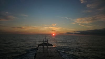 An overhead, wide-angle shot captures a sunset over a dark, choppy sea. The sun, a bright orange orb, is positioned just above the horizon line, casting a warm glow across the sky and reflecting on the water. The sky transitions from a deep blue at the top to shades of orange and pink near the horizon, with wispy clouds streaking across. In the foreground, the deck of a ship is visible, with a metal railing and what appears to be a mast structure. The ship is moving, creating small waves and white foam in its wake. There are no people visible in the frame. The image is likely taken from a ship at sea.