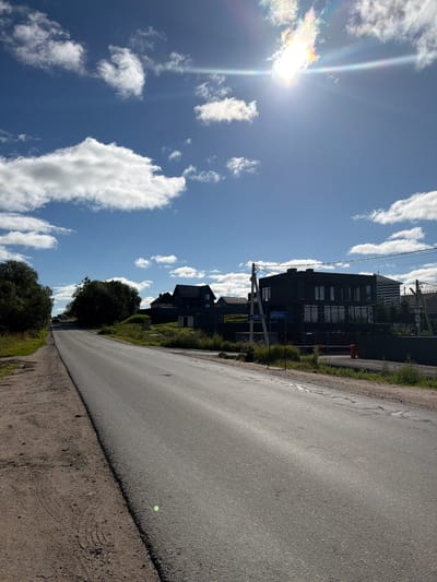The image depicts a roadside view in Leskolovo, Russia, on a sunny day with a bright sun and scattered clouds. The road is paved and extends into the distance, with a dirt shoulder on the left side. Several modern, dark-colored houses are visible on the right side of the road, set back from the street behind fences and greenery. One house is particularly prominent, featuring large windows and a flat roof. A street sign with Cyrillic text is mounted on a utility pole near this house. Trees and vegetation line the sides of the road and the properties. In the distance, more houses and trees are visible, suggesting a suburban or semi-rural residential area. There are no people or vehicles immediately apparent on the road in the foreground. The sun is high in the sky, creating a lens flare effect.