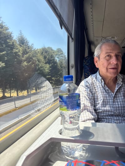 The image shows a man in a bus in San Miguel Topilejo, Mexico. The man is elderly with gray hair and is wearing a plaid shirt. He is sitting next to a window with a reflective view of trees and a road. A bottle of water is on a table next to him. The bus interior is visible, including the window shade, seat, and overhead compartments.