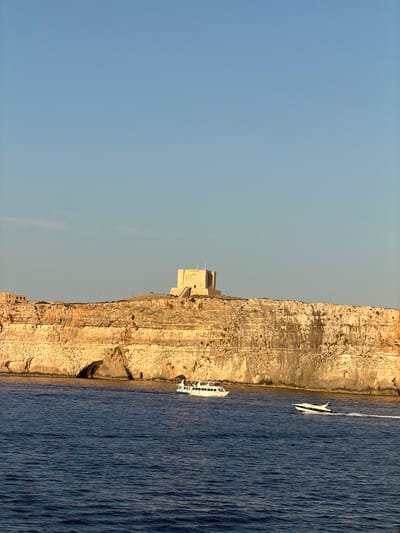 The image captures a serene coastal scene with a prominent cliff face overlooking a deep blue sea. Perched atop the cliff is a large, rectangular stone structure, appearing to be a historical fortification or watchtower. Two boats are visible on the water: a larger, white passenger boat with multiple people on deck, sailing closer to the cliff, and a smaller, faster white speedboat creating a wake as it moves to the right of the frame. The cliff itself is characterized by its rugged, layered texture, with visible caves and overhangs at its base. The sky above is a clear, pale blue, indicating fair weather. The overall impression is one of a tranquil day at the coast, with the ancient structure serving as a focal point against the natural landscape. This is likely a view of the coastal area near Għajnsielem, Malta, given the distinctive architecture and natural formations often found in the region.
