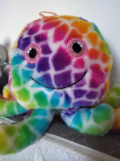 A plush toy octopus with a rainbow-colored, spotted pattern is positioned on a surface. The toy has two large, circular pink eyes with black pupils and a smiling mouth. It has a loop on the top of its head. The octopus is on what appears to be a striped fabric surface, with another gray textured object to its left. The background is a plain white wall. The image was taken in Darlington, United Kingdom.