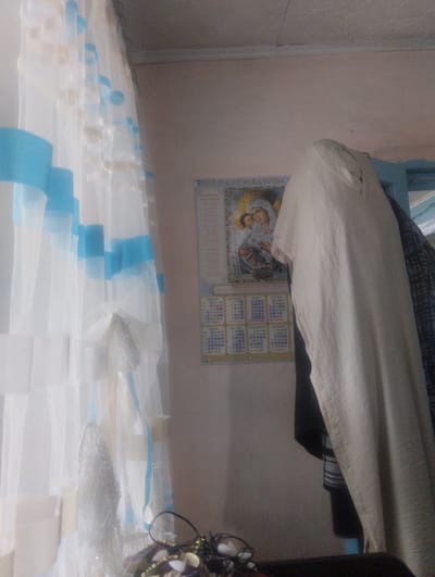The image appears to be an indoor scene, possibly a residential room, in Poltavka, Kyrgyzstan. On the left side of the frame, a sheer white curtain with blue and beige stripes is prominently displayed, obscuring a portion of the background. To the right of the curtain, a wall is visible, adorned with a calendar that features an image of the Virgin Mary and child. The calendar also contains text and a grid of dates, suggesting it is a religious or informational calendar.

Hanging on the wall or on a nearby fixture to the right of the calendar is a light-colored, long-sleeved garment, possibly a dress or a robe. It is made of a textured fabric and has buttons along the front. Partially visible behind this garment is another piece of clothing, appearing to be dark with a striped pattern.

In the lower right corner of the image, a dark surface is visible, possibly a table or furniture. On this surface, a small arrangement of decorative items, possibly part of a religious icon or a craft project, can be seen. These items appear to include dark beads, a metallic object, and possibly some feathers or fabric. The overall lighting in the scene is dim, suggesting it might be taken indoors with limited natural light. The image quality is grainy, indicating it may have been taken with a low-resolution camera or in poor lighting conditions.