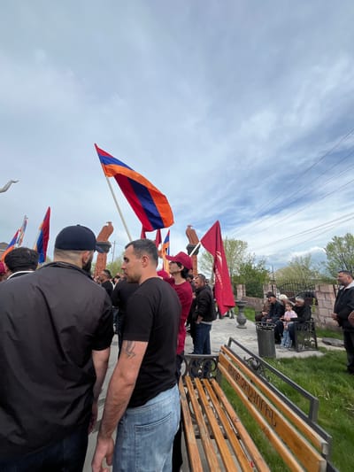 A ground-level, long-shot image captured in Akhuryan, Armenia. The scene shows a group of people standing in front of a low wall or structure with benches, possibly in a public space or memorial. Some of the people are holding Armenian flags and red flags. The group appears to be observing a somber event. There's a cloudy sky overhead, and power lines are visible. In the background, there are hints of other buildings and greenery.