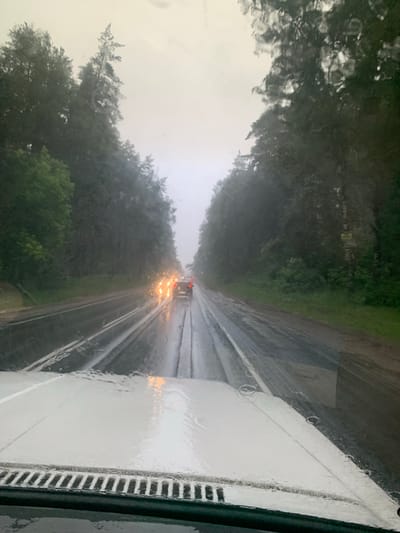 This is a low-angle, outdoor, full shot of a wet road in Zolotovo (Sitnikovskii S/S), Russia, during rainfall. The image is captured from inside a vehicle, looking forward through the windshield, which is streaked with raindrops. The road is dark and reflective, with two visible lanes marked by white lines. Several cars are ahead, their taillights glowing orange through the rain. The road is bordered by a dense forest of dark green trees on both sides. The sky is overcast and light gray, typical of rainy weather. The foreground shows the hood of the vehicle and its windshield wipers, also wet from the rain.