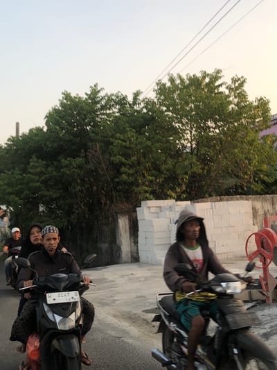 In Sentul, Indonesia, on a paved road during daylight, three people ride a black motorcycle. The driver, a man wearing a black jacket and a patterned skullcap, is in the center. Behind him, a woman in a black headscarf is seated. To their left, a person wearing a white cap rides another motorcycle. In the foreground, a man in a hooded jacket and green shorts rides a separate black motorcycle, looking to the left. The background features a wall constructed of white bricks and lush green trees. A red construction mixer is visible to the right of the brick wall. The sky is a pale blue with some hazy sunlight.