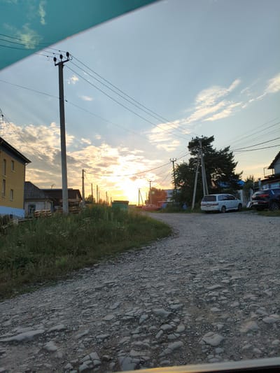 The image depicts a rural street scene in the village of Andreevka, Russia, during sunset. A gravel road curves through the scene, bordered by overgrown grass and vegetation on the left. Several houses are visible, including a yellow building with a blue base on the left and other smaller structures with weathered roofs. Utility poles with overhead power lines are prominent features, with one large pole situated near the center of the frame.

In the background, the sun is setting, casting a warm glow and visible rays through scattered clouds. Several vehicles are present: a white minivan is parked on the right side of the road, and a dark-colored SUV is parked behind it. Further down the road, a red car is visible. The overall atmosphere is one of a quiet, rural evening.