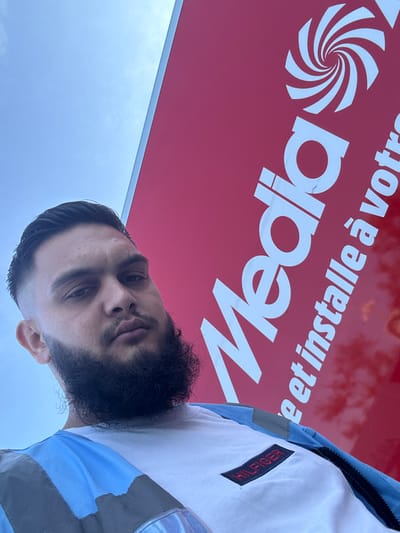 A man with dark hair, a beard, and a fair complexion is captured in a low-angle, selfie-style photograph. He is wearing a white t-shirt with a rectangular red and black patch that reads "HILFIGER" on the left chest area, and a blue and grey jacket. The man is positioned in front of a large, vibrant red sign with white text and a swirling white logo. The sign appears to be advertising a company named "Media" and also includes the text "e et installe à votre". The sky is visible above the man and the sign, appearing light blue with some faint cloud formations. The overall lighting suggests it is daytime. The location is specified as the city of Boom, Belgium.