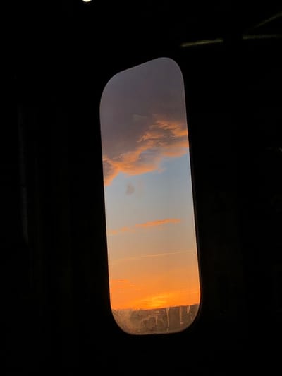 An elongated, slightly blurry rectangular window shows a dramatic sky. Light blue transitions to orange and gold at the right, possibly indicating sunrise or sunset. Dark, textured clouds occupy the left portion of the frame. The rest of the image surrounding the window is in deep shadow, revealing parts of a frame that could belong to a car’s roof or some other dark interior space. The window glass appears dirty or scratched, adding to the atmosphere.