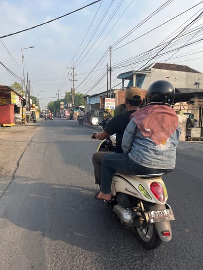 In Kedungkendo, Indonesia, two individuals are riding a light-colored Honda Scoopy motorcycle along a paved road. The rider, wearing a brown cap and a black t-shirt, is in the front. The passenger behind, wearing a pink hijab, a denim jacket with a floral design, and jeans, is seated at the back. The motorcycle has a license plate that reads "L 3733 KT 04-26". The street is lined with small businesses and residential buildings. Overhead, a dense network of power lines crisscrosses the sky. The sun casts shadows on the road, suggesting it is either early morning or late afternoon. The overall atmosphere is that of a typical day in a bustling Indonesian town.