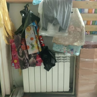 The image depicts a cluttered interior scene, likely in a residential setting in Balakhna, Russia. A white radiator is prominent in the foreground, with various items of clothing and textiles hanging above it. To the left, a sheer yellow curtain is visible. To the right of the radiator, a stack of cardboard boxes wrapped in plastic film is present. The background shows wooden shelving filled with colorful items, possibly rolls of fabric or other stored goods. The overall impression is one of a disorganized storage area. There are no people or animals visible in the image.