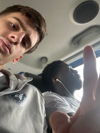 The image depicts two males inside a passenger vehicle, likely a bus or large van. The male in the foreground, positioned left, has light skin, brown hair, and looks directly at the camera with a neutral expression and slightly pouted lips. He wears a light grey zippered jacket featuring a circular emblem on the left chest that reads "STRASBOURG FC 67" and "PLUS QU'UNE VILLE, UNE FIERTE". A finger from his right hand is prominently visible in the bottom right corner, partially obscuring the second individual. The second male, situated to the right and slightly behind, has dark skin, short dark curly hair, and a beard. He is seen looking upwards and to the right, away from the camera, wearing a light grey t-shirt and white wired earphones. The vehicle's interior features a light-colored ceiling with a dark circular speaker on the right and a light-colored grab handle. A blue-tinted window is visible on the far right. While no specific visual cues within the image confirm the precise location, the "STRASBOURG FC 67" emblem suggests an association with the Strasbourg area in the Bas-Rhin department (67), where Bernolsheim, France, is located.