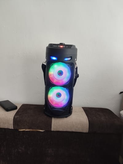 A black, upright portable speaker with two illuminated, multi-colored LED speaker cones is centered on a dark brown and tan upholstered surface. The speaker has a rugged, angular design with blue LED accent lights above the speaker cones, resembling eyes. A black strap is attached to the top of the speaker.

To the left of the speaker, on the same surface, rests a black rectangular electronic device, possibly a power bank or phone. The surface appears to be the top of a bench or storage ottoman, with a darker brown fabric below a lighter tan band.

The background is a plain, textured white wall, with some visible imperfections and marks. The lighting suggests an indoor setting, likely during the daytime due to the ambient light. No people are visible in the image.

Visible text on the speaker includes "ZQ54239" and possibly a small logo near a red indicator light at the top. The overall impression is of a modern electronic device set up for use or display in a home environment. The illuminated speaker cones suggest the speaker is either playing music or has its lighting feature activated.