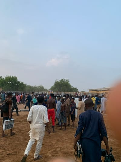 The image depicts a large crowd of people gathered in an open space. The location is Sokoto, Nigeria, specifically at latitude 13.049 and longitude 5.238. The ground is dusty and uneven, suggesting a dry and possibly arid climate. The sky is a light blue, indicating a clear day or early evening. There are trees in the background and a low-lying building, suggesting a suburban or rural setting. The people in the crowd are mostly men and boys, dressed in various styles of clothing. Some appear to be engaged in conversation or watching something in the distance. Others are standing around, seemingly waiting or observing. In the foreground, a man in a white robe walks towards the camera, holding a white case. Behind him, a man in a dark blue shirt holds a black camera and a microphone. The overall scene suggests a casual gathering of people, possibly for a social event, religious event or an activity.