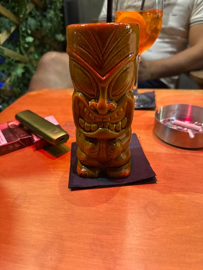 The image captures a close-up, eye-level view of a tiki mug positioned on a dark napkin atop an orange wooden table. To the left of the tiki mug, a green e-cigarette and a box of cigarettes are visible. In the background, a glass filled with an orange beverage and an orange slice, accompanied by a straw, is slightly out of focus. An ashtray containing cigarette butts is to the right of the tiki mug. The scene appears to be set in an outdoor seating area of a bar or restaurant in Chersonisos Municipal Unit, Greece, suggested by the lush greenery and ambient lighting. The overall impression is one of a relaxed, evening setting.