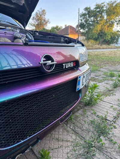 The image shows the front of a modified car with a vibrant purple and teal iridescent paint job. The hood is open, revealing engine components and blue hoses. A chrome Opel emblem is visible on the grille, and a "TURBO" badge is also present. A Bulgarian license plate, reading "C 0380 BG", is attached to the front bumper. The car is parked on a cracked concrete surface overgrown with weeds, in what appears to be a residential area with houses and trees in the background. The lighting suggests it is late afternoon or early evening. The location is identified as Lepitsa, Bulgaria.