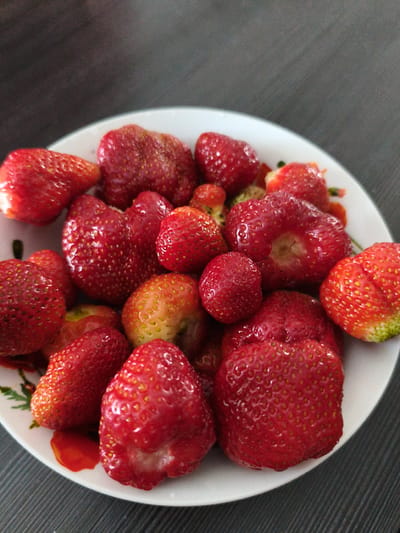 A close-up, overhead shot features a white, circular plate filled with a variety of fresh strawberries. The strawberries are predominantly bright red, with some exhibiting lighter red and yellow hues. A few strawberries show visible green tips, indicating they are nearing ripeness or are just past their peak. The plate has a subtle floral pattern along its rim. The strawberries are arranged densely, with some overlapping. The background consists of a dark, textured surface, possibly a wooden table, with a subtle sheen. The lighting is even, highlighting the glossy texture of the strawberries. This image was taken indoors.